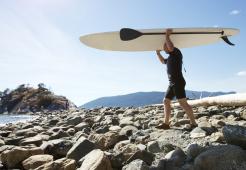 Mature adult carrying paddle board across the beach towards the sea.